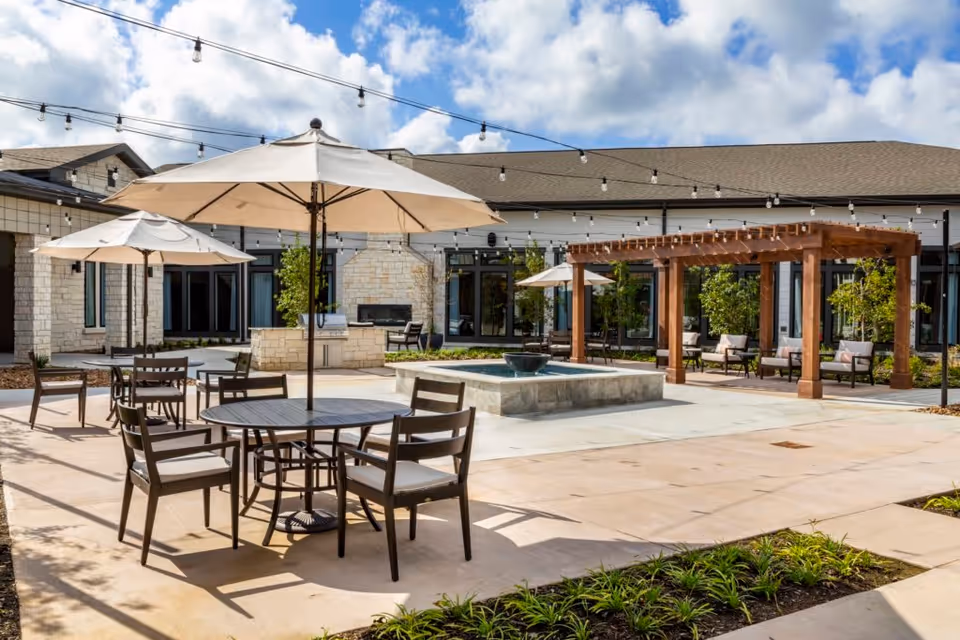 Outdoor courtyard area with round tables and chairs under large beige umbrellas, a central water fountain, string lights overhead, a wooden pergola with seating underneath, and a stone building in the background under a partly cloudy sky.