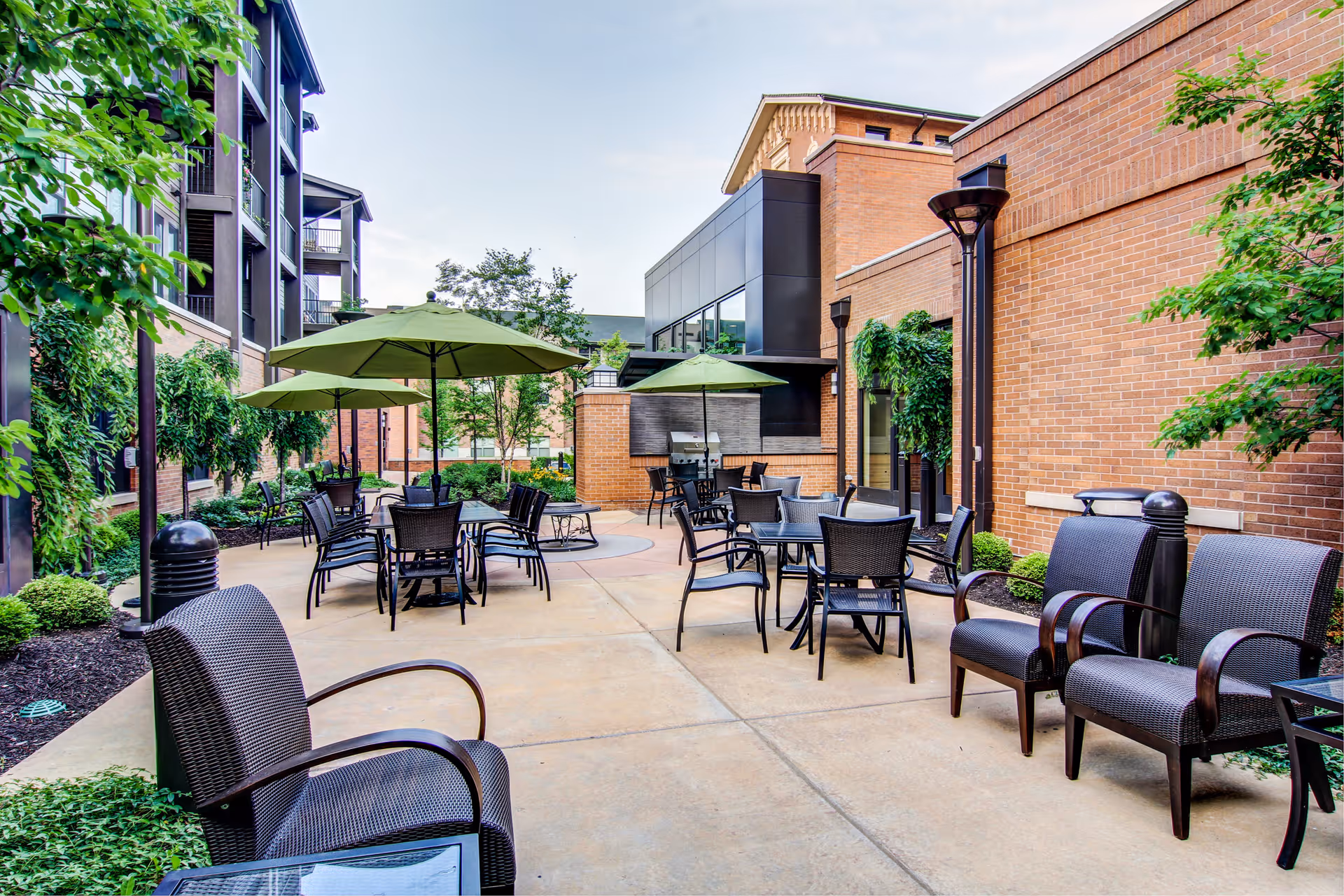 Outdoor patio area at EverTrue Laclede Groves featuring multiple tables with green umbrellas, black chairs, and lounge seating. The space is surrounded by brick buildings and greenery, with a clear sky above.