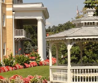 Outdoor view of a senior living facility showing a white gazebo surrounded by green grass and colorful flower beds, with part of a brick building and a balcony visible in the background under a clear sky.