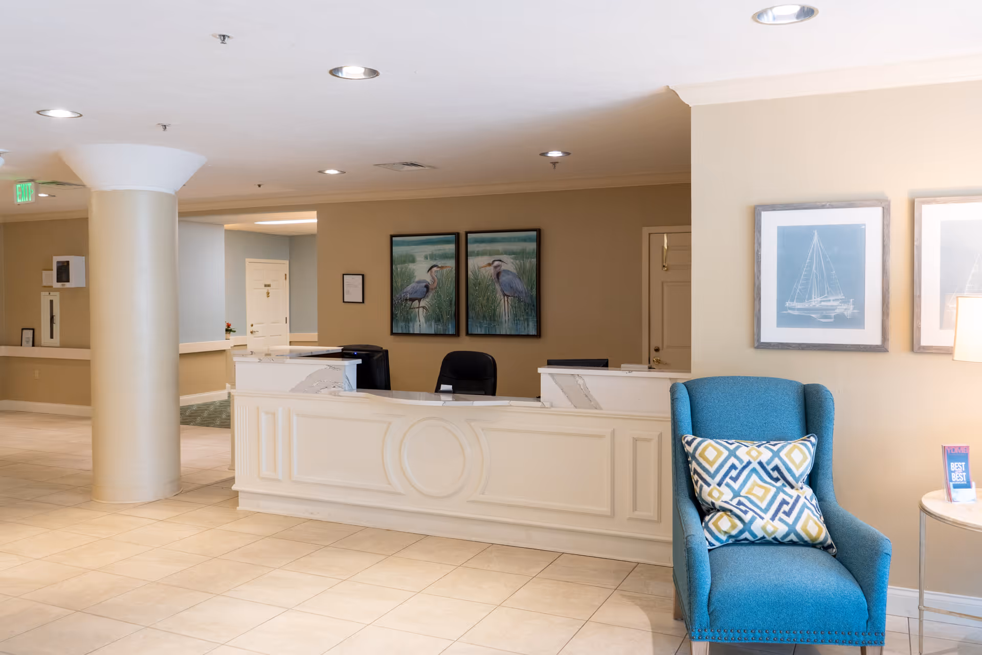 Reception area of Heron Club at Prestancia featuring a white front desk with two computer monitors, a black office chair, beige walls adorned with two framed paintings of herons, a blue upholstered armchair with patterned cushions, and two framed pictures of sailboats on the wall beside the chair.