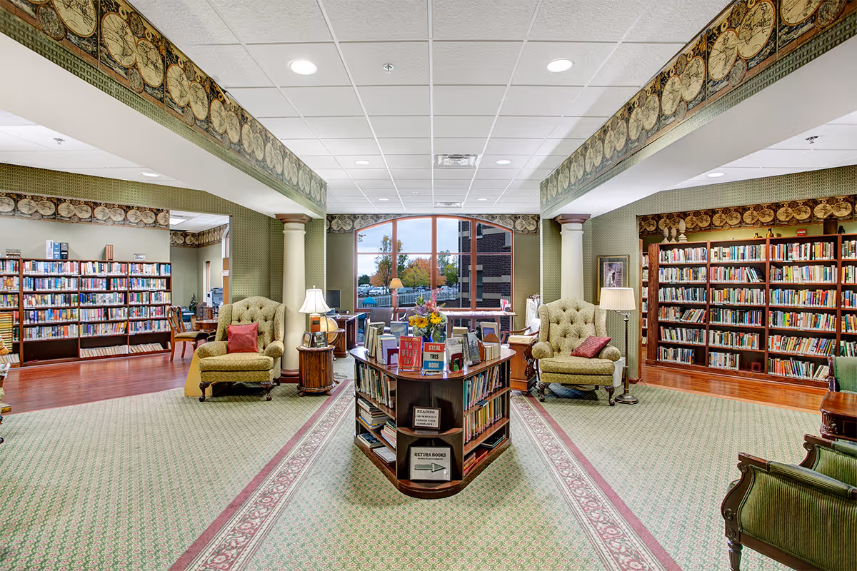 A spacious and well-lit library room with green patterned carpet and wallpaper. The room features two large armchairs with red cushions, a central wooden book display filled with books, and bookshelves lining the walls. A large window at the back lets in natural light and shows an outdoor view with trees and a building. The ceiling has recessed lighting and decorative molding along the top edges of the walls.