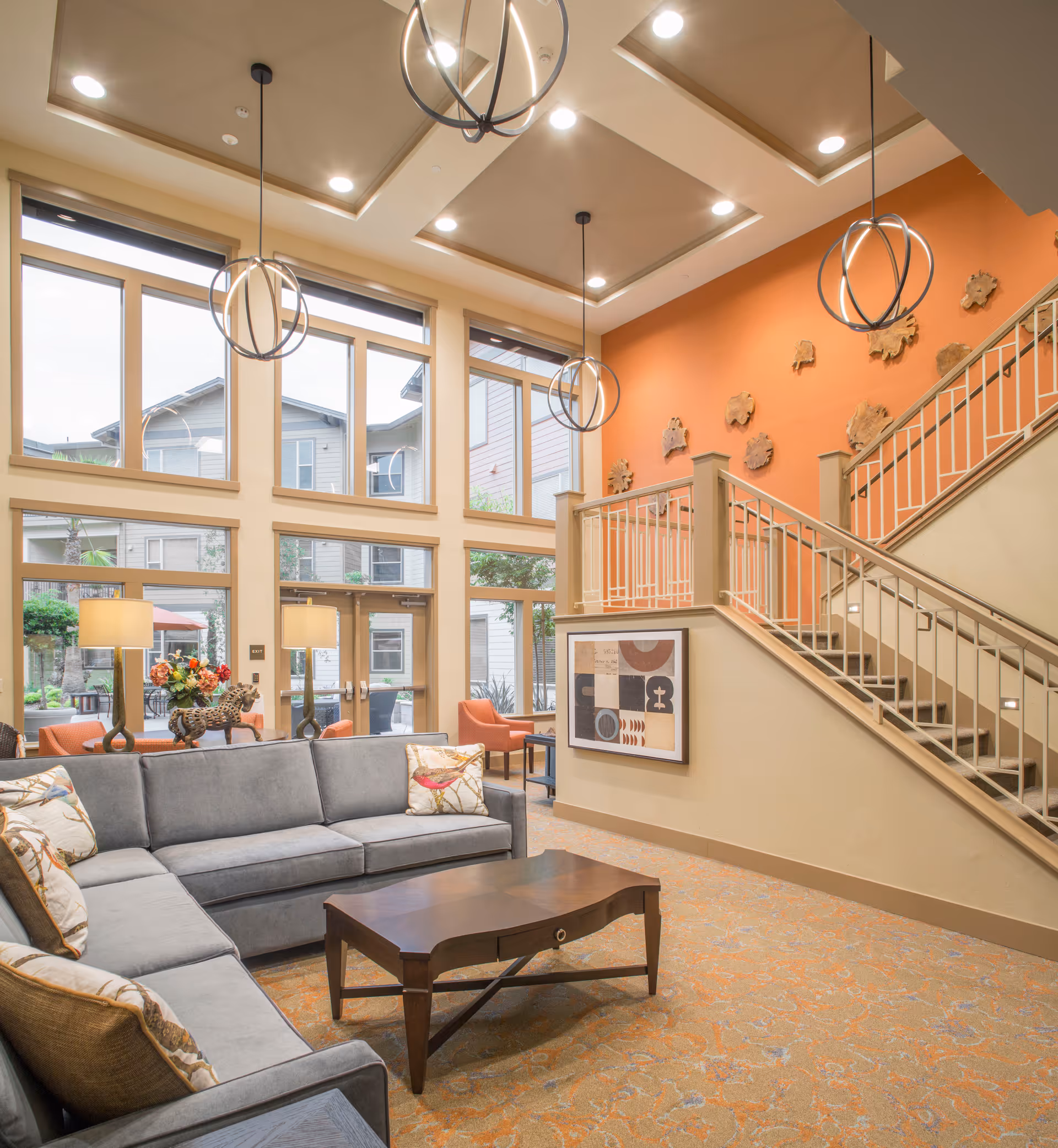 A spacious and well-lit living room area with large windows allowing natural light to fill the space. The room features a gray sectional sofa with decorative pillows, a wooden coffee table, and modern pendant lights hanging from a recessed ceiling. There is an orange accent wall with artistic wall decorations and a staircase with a beige railing. Outside the windows, parts of the building and greenery are visible.