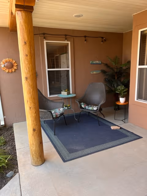 Covered porch with two wicker chairs and a small table on a rug beside a wooden support post and potted plants.