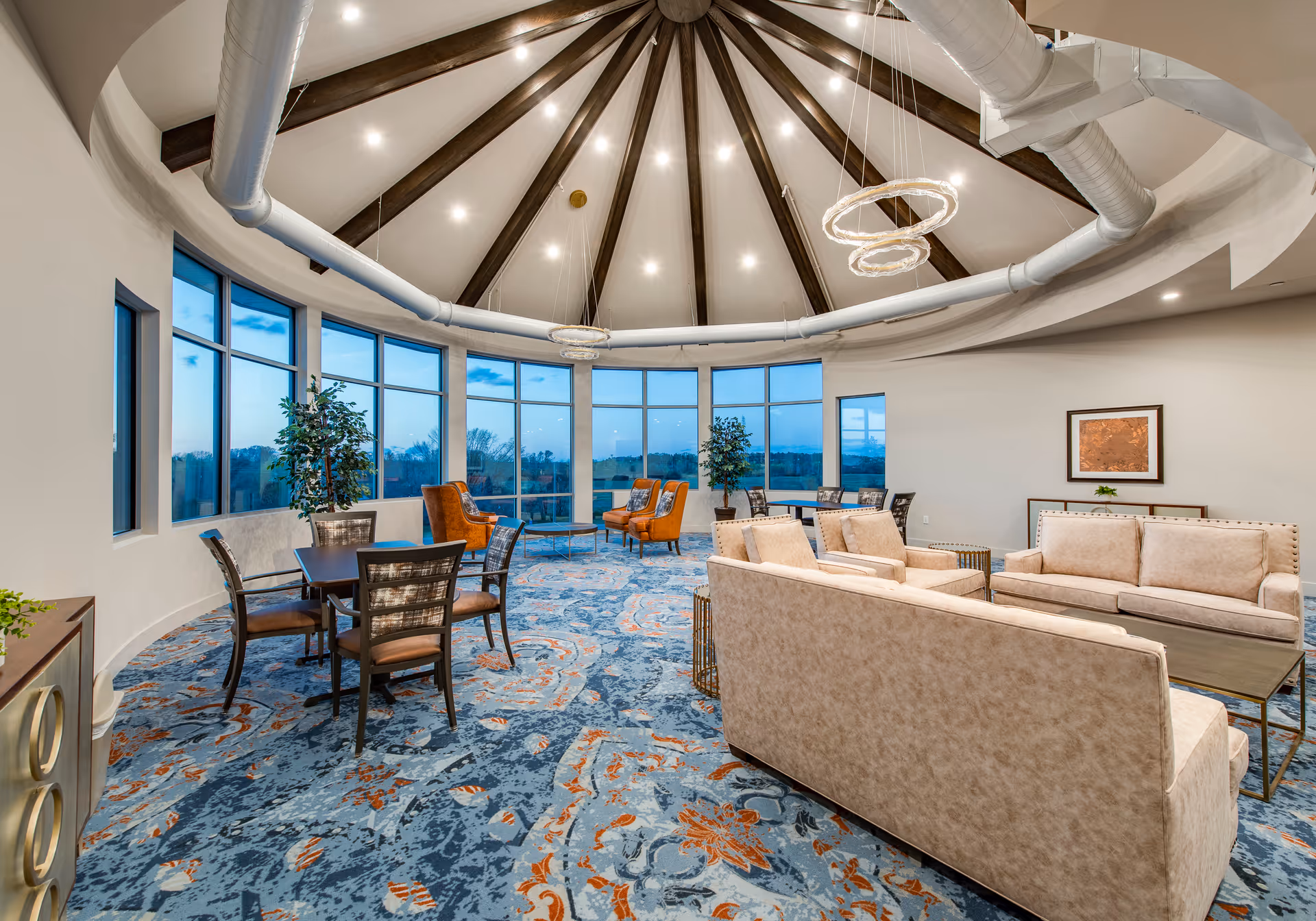 A spacious, circular lounge area with large floor-to-ceiling windows offering a scenic view outside. The room features a high ceiling with exposed beams and modern circular light fixtures. There are beige sofas, armchairs, and several tables with chairs arranged on a patterned blue and orange carpet. Potted plants and framed artwork decorate the space.
