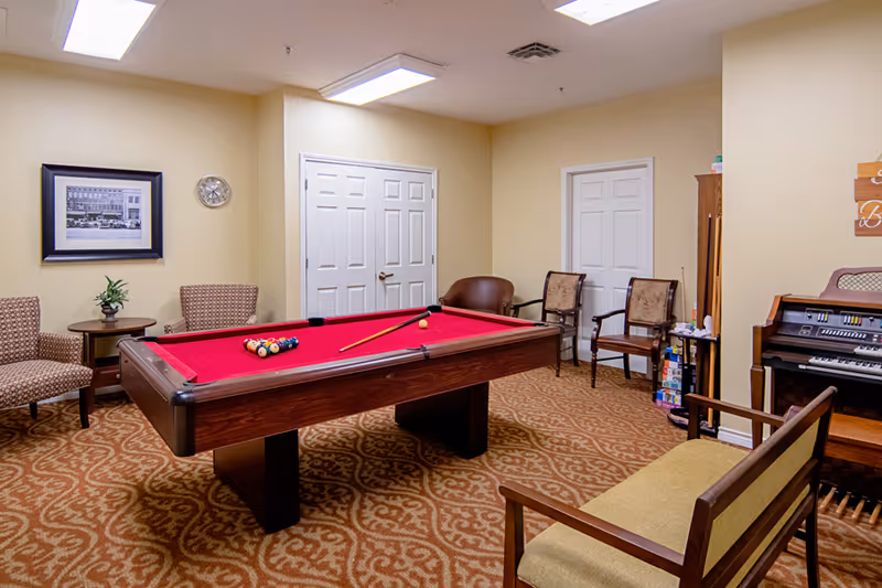 A cozy game room featuring a red felt pool table with billiard balls and a cue stick arranged on it. The room has patterned carpet flooring, beige walls, and several chairs including armchairs and a bench. There is a framed black and white photo and a clock on one wall, and an organ with sheet music on the right side. Two white doors are visible in the background.