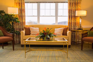 Well-lit living room with a beige sofa, glass coffee table holding a floral arrangement, two armchairs, side lamps, and a large window behind.