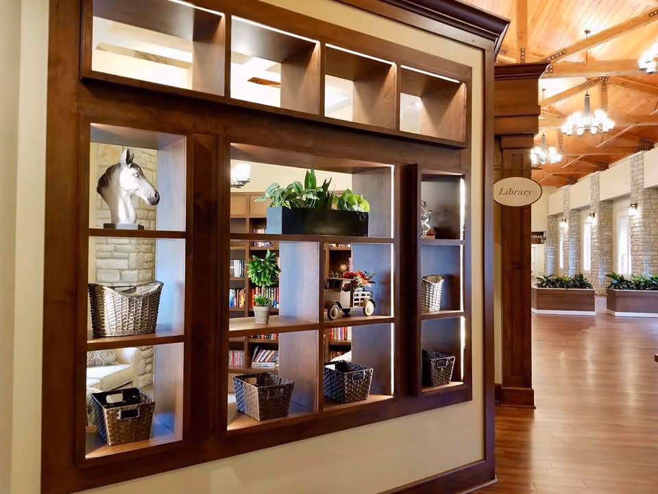 Interior view of a senior living facility featuring a wooden shelving unit with decorative items including a horse head sculpture, plants, and baskets. In the background, there is a hallway with wooden flooring, stone walls, and a sign indicating the library. The ceiling has wooden beams and hanging light fixtures.