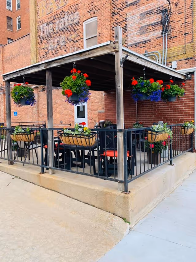 Outdoor patio area with a wooden pergola structure, black metal railing, and several hanging flower baskets with red and purple flowers. There are tables and chairs with red cushions underneath the pergola. The background shows a brick building with faded painted signage.