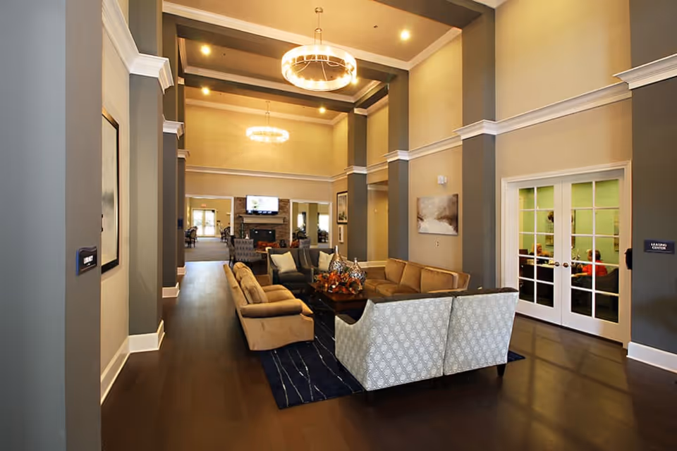 Spacious communal lounge with high ceilings and chandeliers, several sofas and chairs arranged around a coffee table, and French doors on the right.