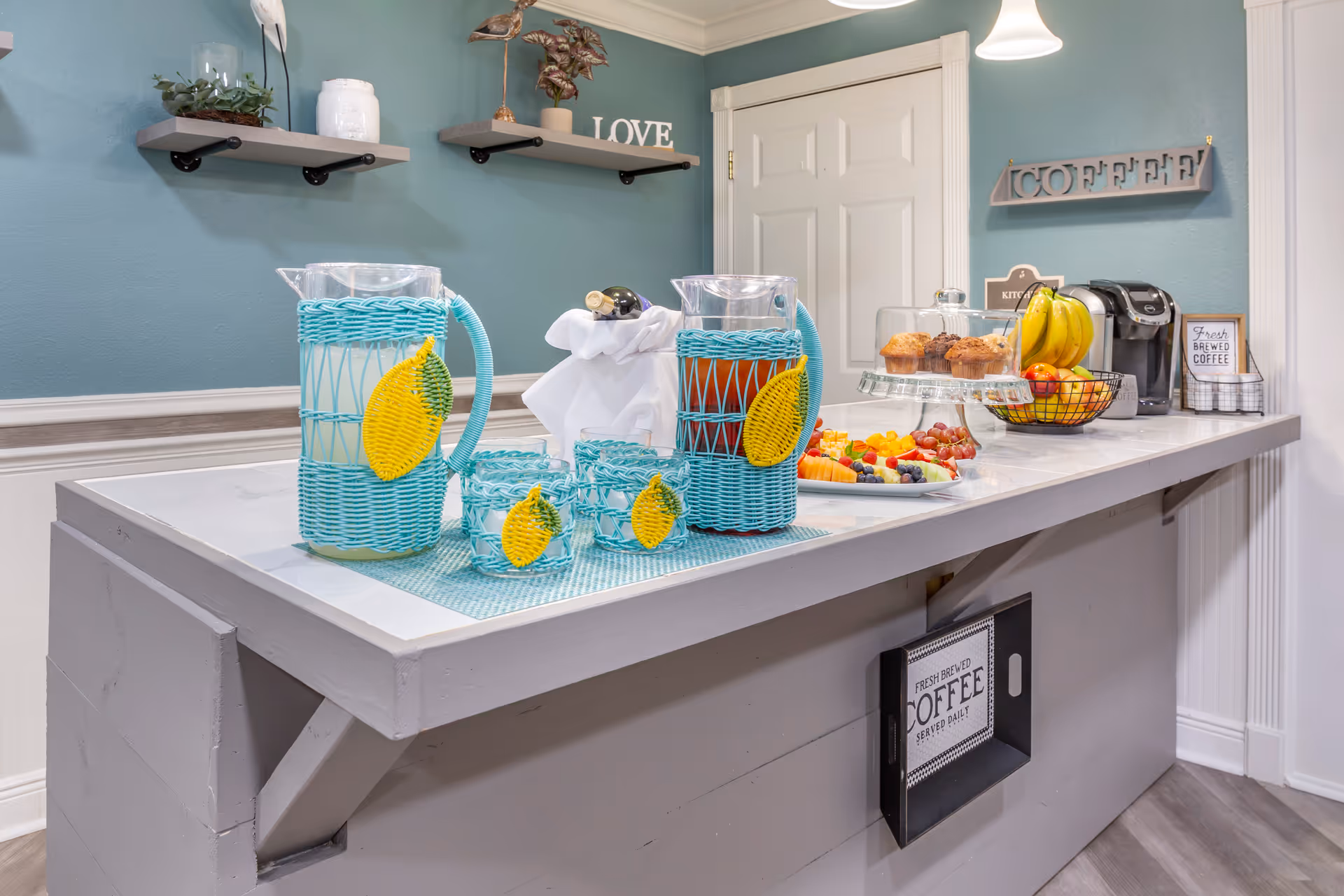 A kitchen counter with two pitchers and four glasses in blue woven holders, a plate of assorted fresh fruit, a glass cake stand with muffins, a basket of bananas and apples, a coffee maker, and decorative signs on the wall including one that says 'COFFEE' and another that says 'Fresh Brewed Coffee'. The wall is painted teal with white trim and two small shelves holding decorative items.