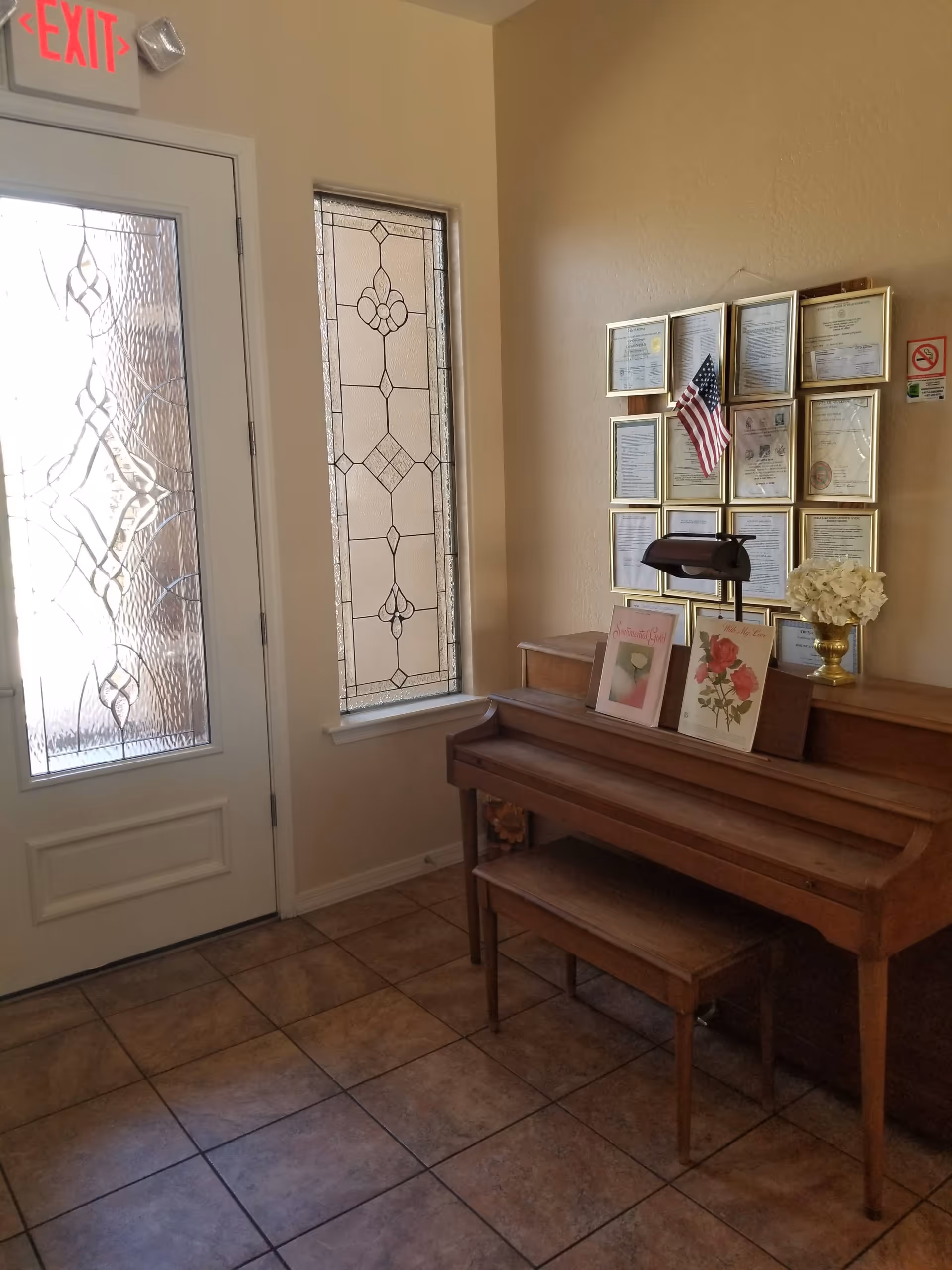 A corner of an interior room with a wooden piano and matching bench. Above the piano are framed certificates and an American flag on the wall. To the left is a door with decorative frosted glass and a stained glass window beside it. The floor is tiled and there is an exit sign above the door.