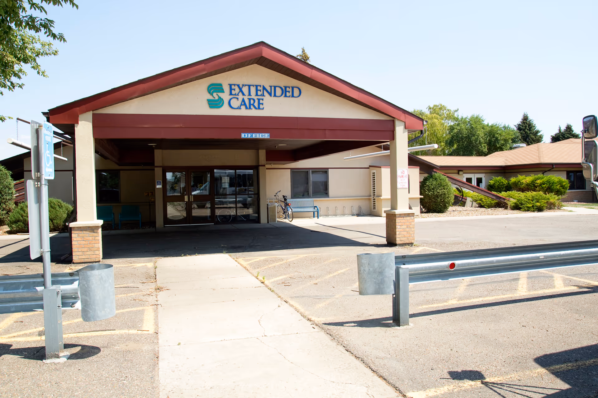 Front exterior view of Sidney Health Center Extended Care building with a covered entrance, wheelchair accessible parking, and surrounding greenery under a clear sky.