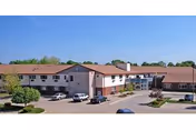 Exterior view of a two-story senior living facility building with a red roof, beige walls, and multiple windows. Several cars are parked in front of the building under a clear blue sky.