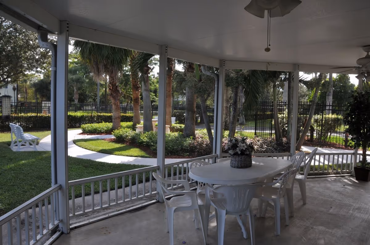 Covered outdoor patio area with a white plastic table and chairs, a flower arrangement on the table, ceiling fans, and a view of a landscaped garden with palm trees, bushes, a curved walkway, and a white bench.