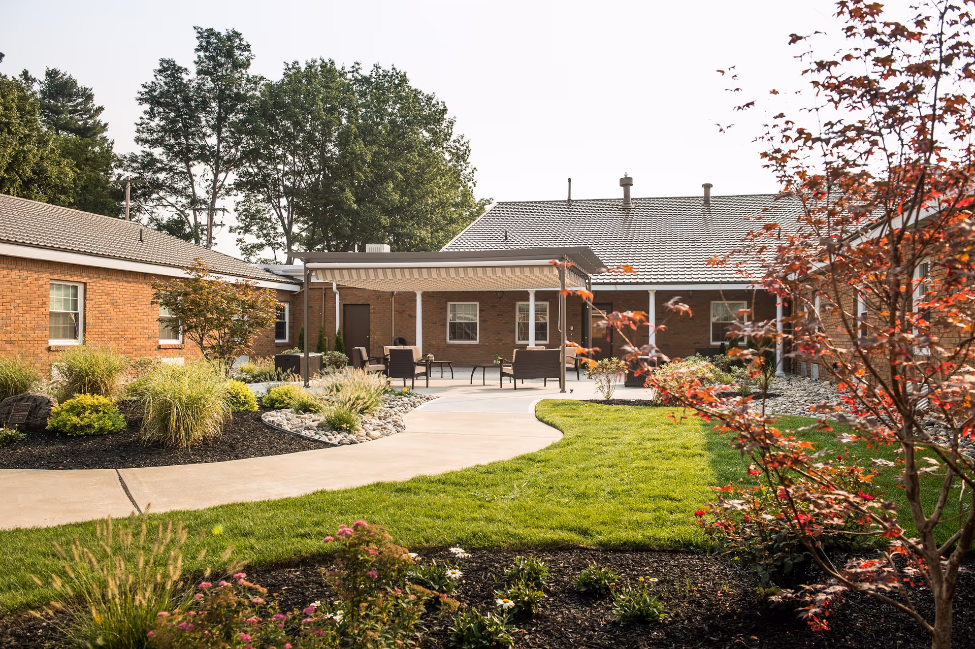 Outdoor courtyard area at Kingsway Arms Nursing Center featuring a paved walkway, green grass, landscaped garden beds with various plants and flowers, and a covered patio with outdoor seating. The surrounding buildings are made of brick with windows and a tiled roof.