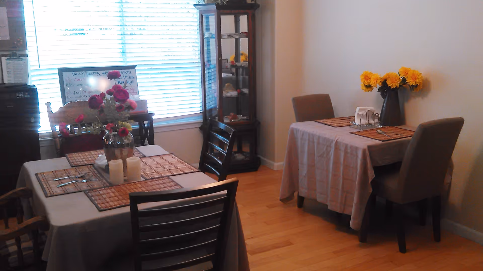 A dining area with two tables covered with tablecloths and set with placemats, utensils, and napkins. One table has a vase with red flowers and candles, while the other has a vase with yellow flowers. There is a wooden chair, a glass display cabinet, and a window with blinds in the background.