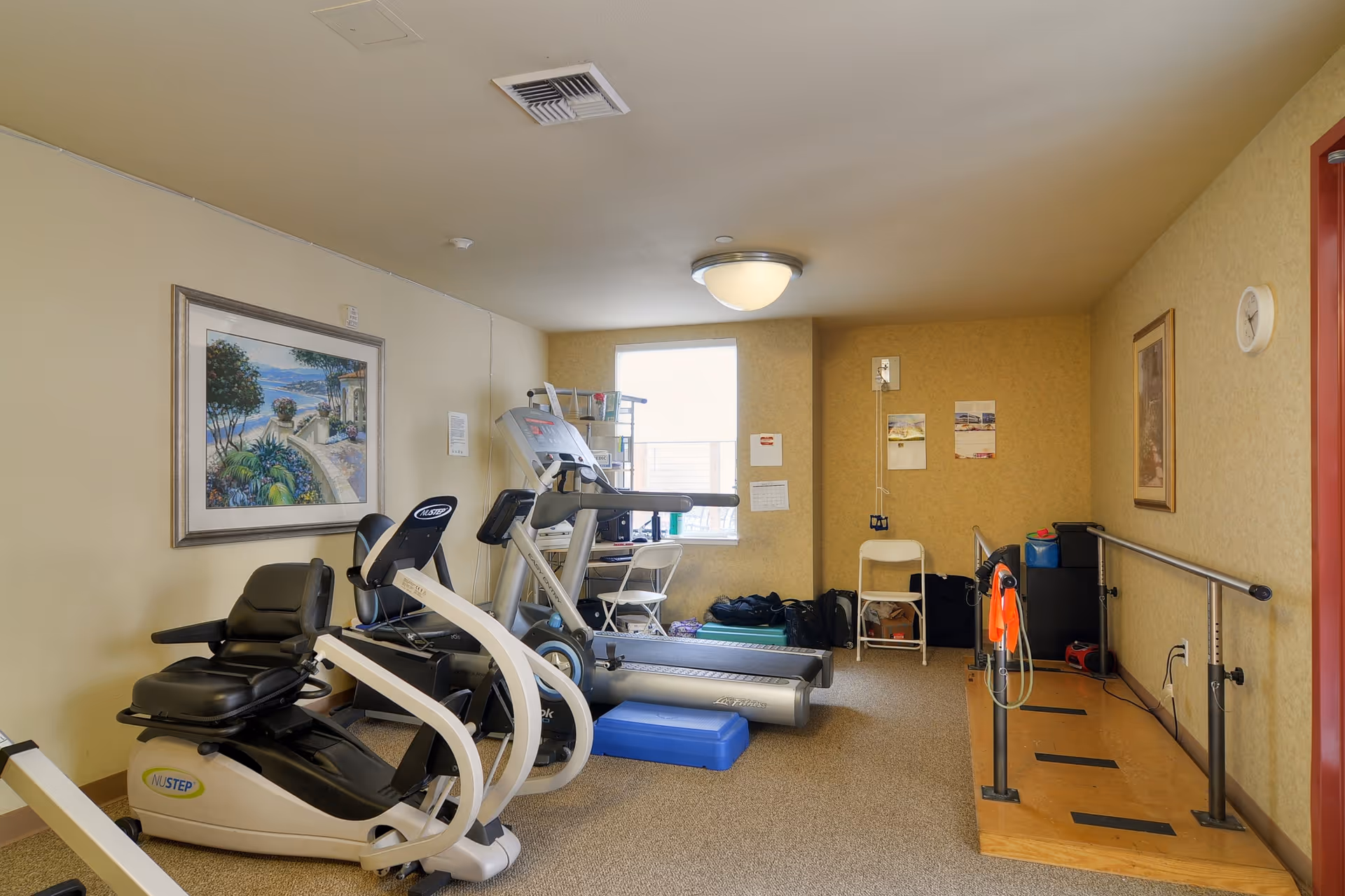A small fitness room with exercise equipment including a recumbent bike, treadmill, and elliptical machine. There is a wooden platform with parallel bars for walking exercises on the right side. The room has beige walls, a window letting in natural light, a framed painting on the left wall, and a ceiling light fixture.