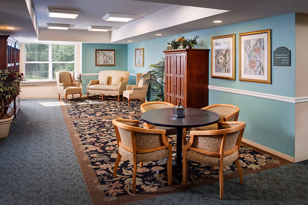 A cozy senior living common area with a round table surrounded by four upholstered chairs in the foreground. In the background, there is a seating area with a loveseat and two armchairs, a large window letting in natural light, a wooden cabinet, potted plants, and framed floral artwork on teal-colored walls.