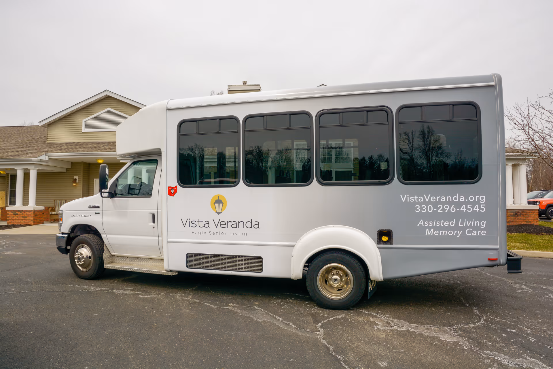 A white and gray shuttle bus parked in front of a senior living facility building. The bus has the Vista Veranda logo and text that reads 'Vista Veranda Eagle Senior Living' and contact information including a website, phone number, and services offered: Assisted Living and Memory Care.