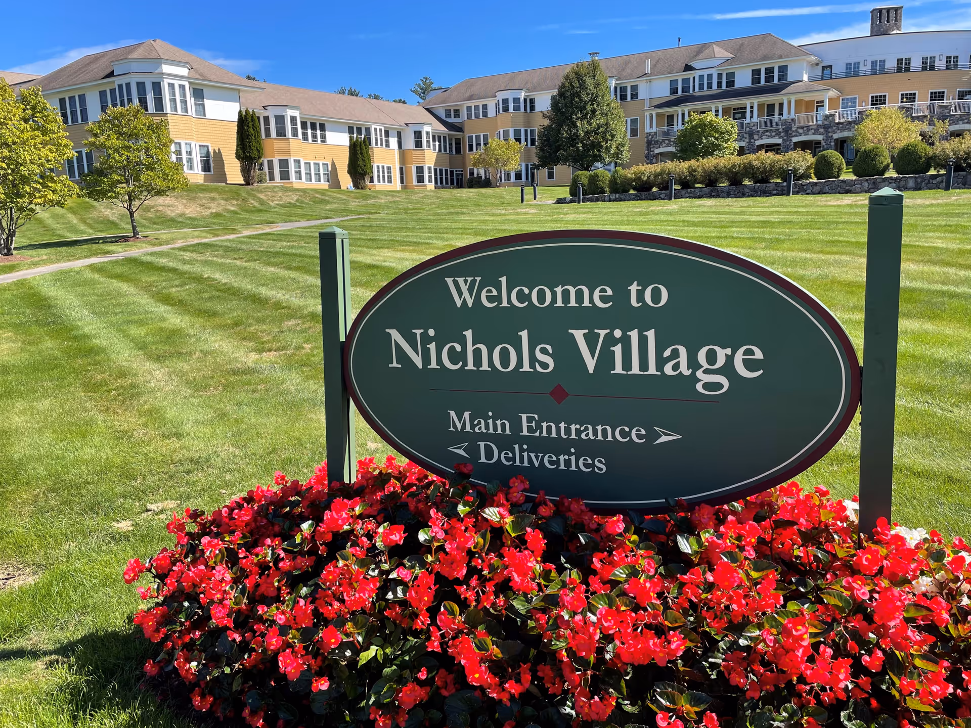 A large green sign with white text reading 'Welcome to Nichols Village Main Entrance Deliveries' is surrounded by vibrant red flowers. Behind the sign is a well-maintained lawn and a large yellow building with white trim and multiple windows under a clear blue sky.