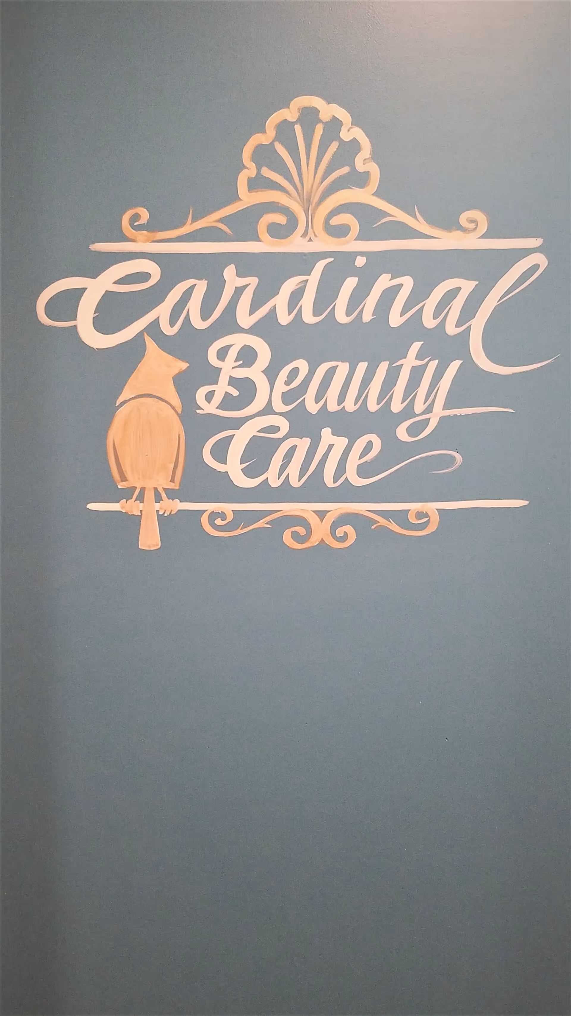 A decorative wall sign with the words 'Cardinal Beauty Care' painted in elegant script on a teal background, featuring a stylized cardinal bird and ornamental flourishes above and below the text.