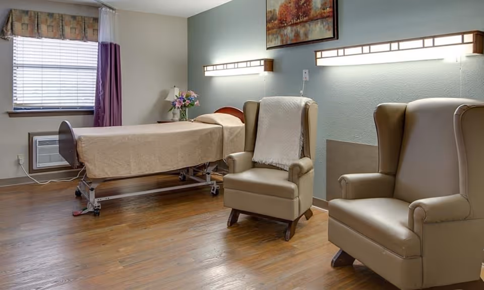 A nursing facility room with a single bed, two upholstered armchairs, wall lights and a window.
