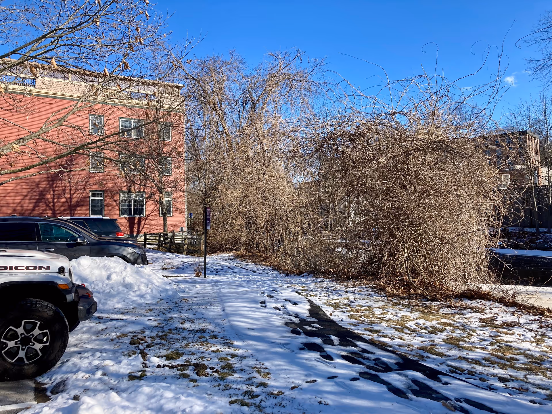 Snow-covered parking area and walkway beside a red multi-story building and leafless trees under a clear blue sky.