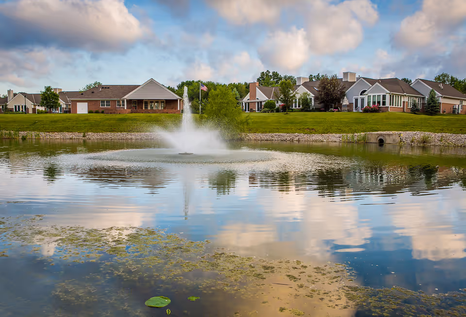 A serene pond with a water fountain in the center, surrounded by well-maintained grass and several single-story residential buildings in the background under a partly cloudy sky.