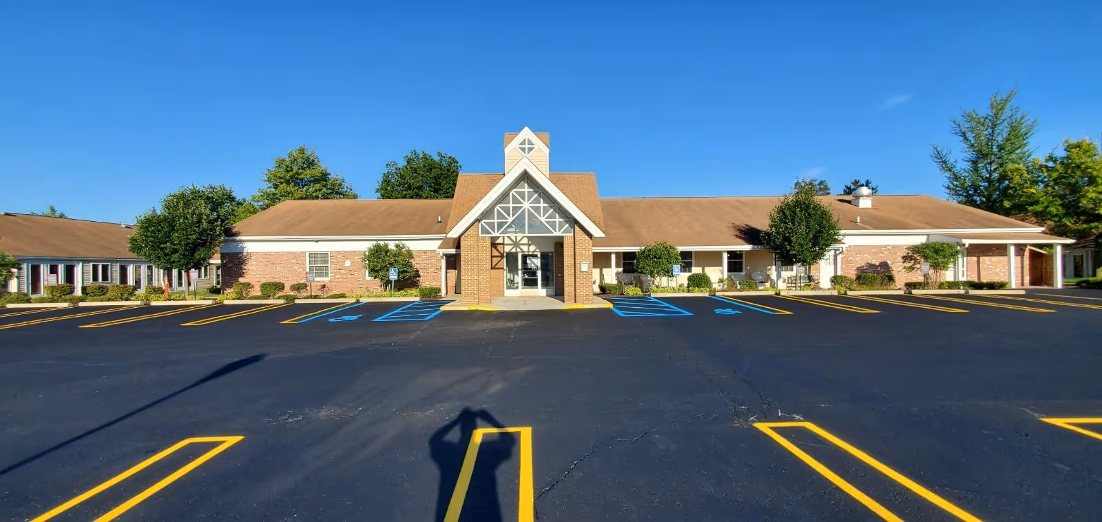Single-story brick senior living facility front entrance with an empty parking lot and marked handicap spaces under a clear blue sky.