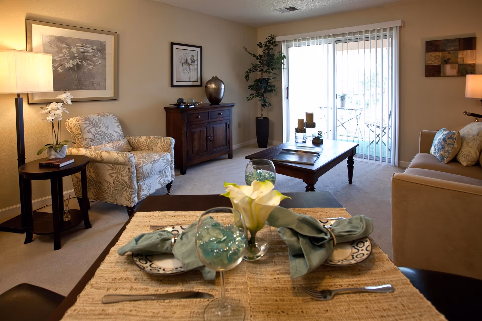 A cozy living room with a beige armchair and a side table with a lamp and a small plant. A wooden cabinet with decorative items is against the wall. A coffee table with candles and small plants is in front of a sliding glass door with vertical blinds leading to a small outdoor patio with a table and chairs. A beige sofa with patterned pillows is partially visible on the right. In the foreground, a dining table is set with plates, green napkins, wine glasses, and a small flower arrangement.