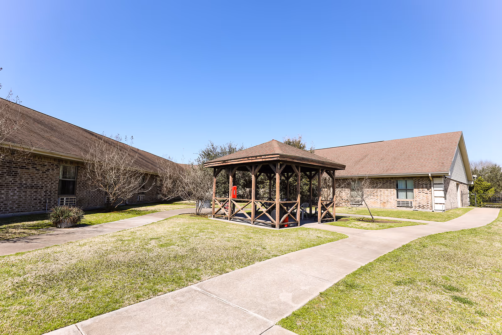 Outdoor area of a senior living facility with a wooden gazebo in the center, surrounded by paved walkways and grassy lawns. Two single-story brick buildings with brown roofs are visible in the background under a clear blue sky.