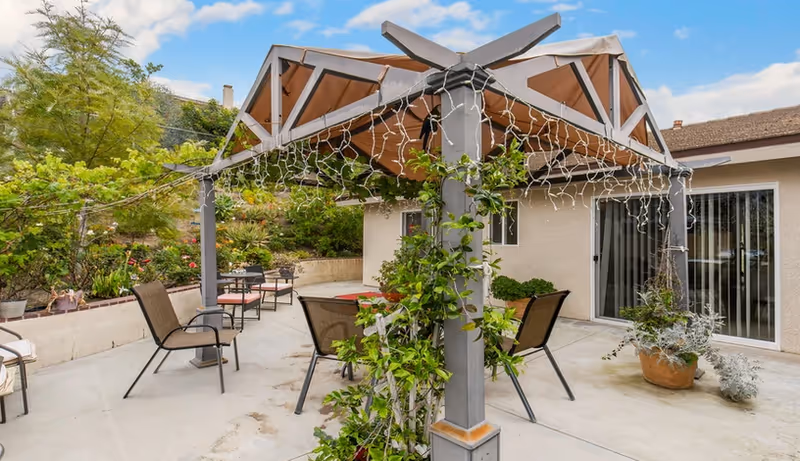 Outdoor patio area with a pergola covered by a brown canopy and decorated with string lights. Several chairs and tables are arranged on the concrete floor. Surrounding the patio are plants and greenery, with a beige building featuring sliding glass doors in the background under a partly cloudy sky.