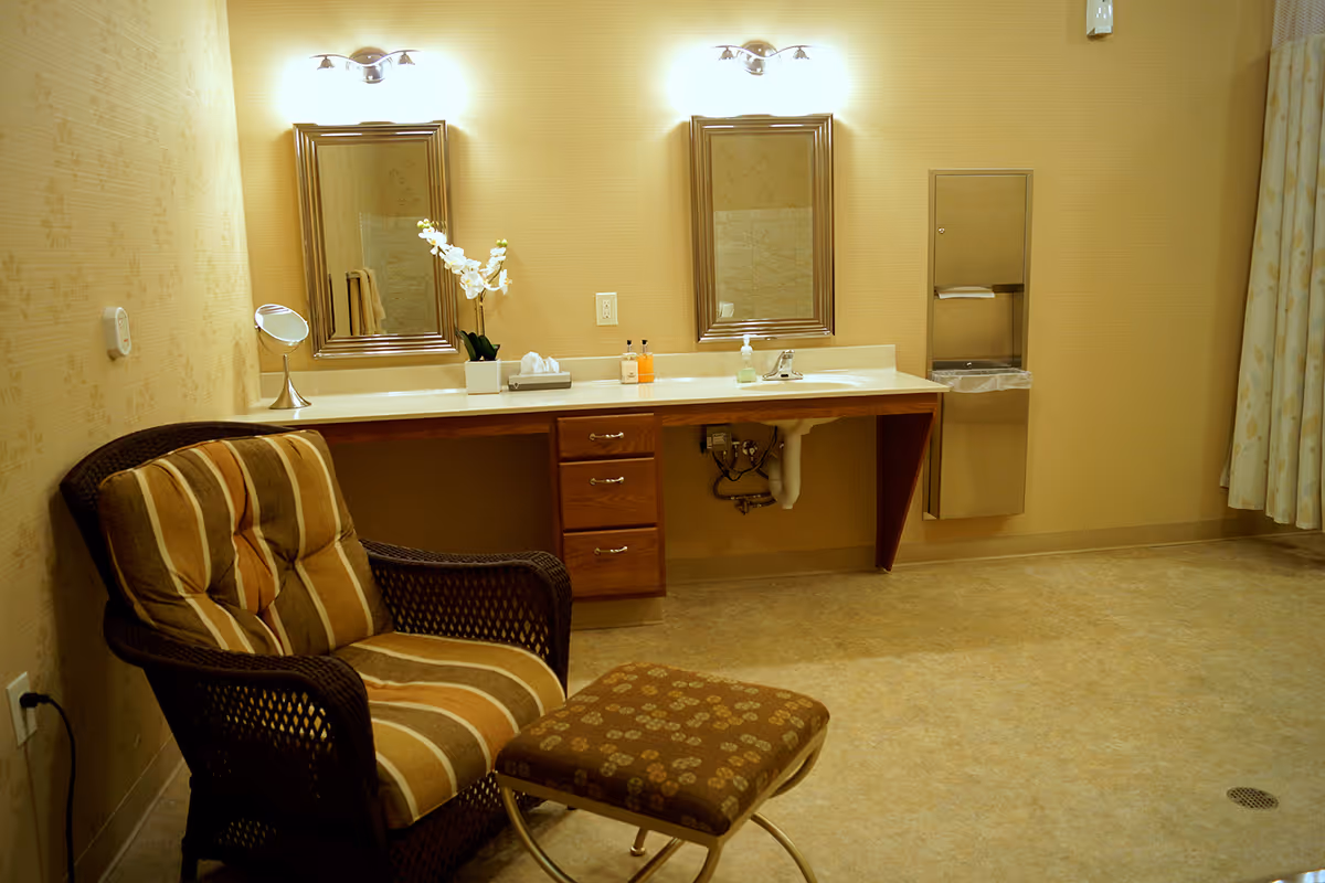 Warm-toned room with a wicker armchair and ottoman in front of a double-sink vanity with mirrors and lights.