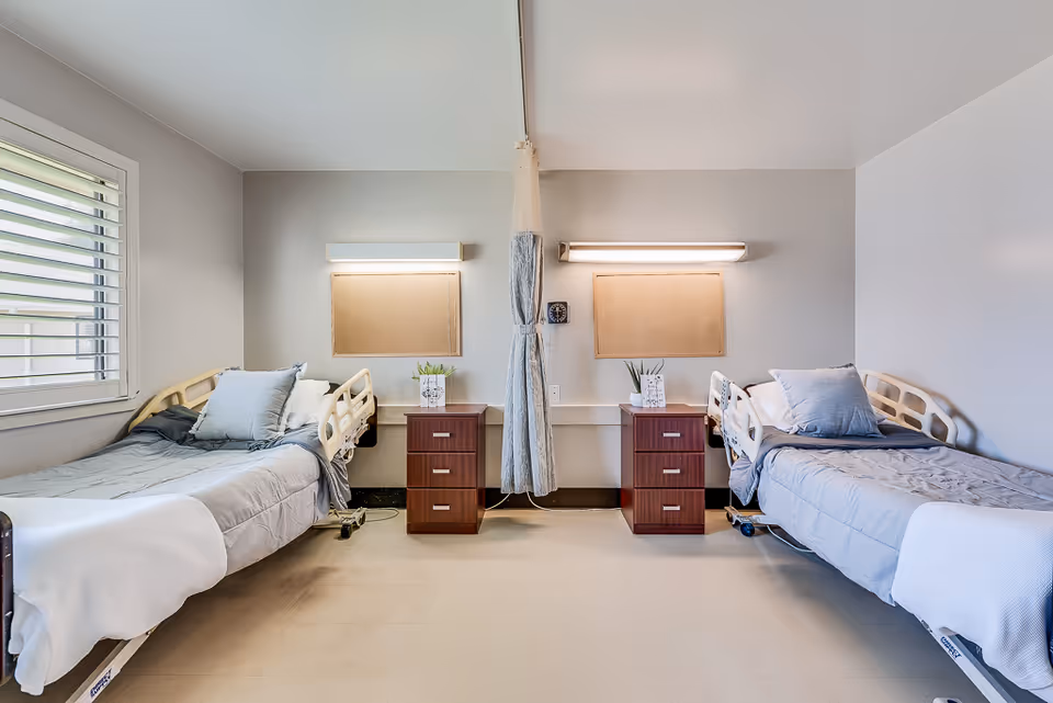 A clean and bright nursing center bedroom with two hospital-style beds separated by a privacy curtain. Each bed has gray bedding and pillows, and there are two wooden nightstands with small plants on top. A window with white blinds is on the left wall, and two rectangular wall lights are above each bed.