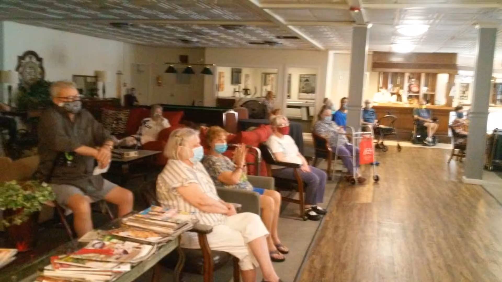A group of elderly people wearing face masks sitting spaced apart in a common area with chairs and sofas. The room has wooden flooring, a bar area in the background, and a table with magazines in the foreground.