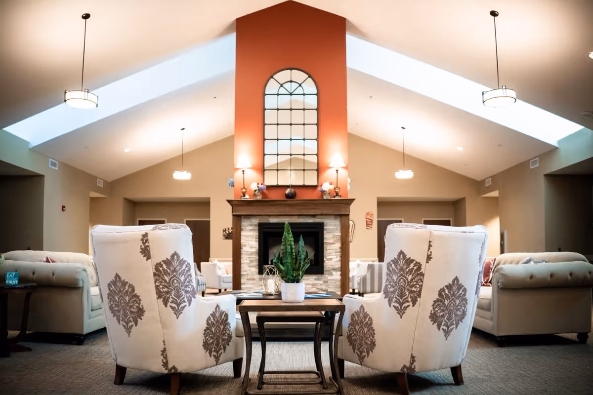A cozy living room area with two patterned armchairs facing a stone fireplace with a tall red chimney. A decorative mirror and lamps are on the mantel. Sofas and additional seating are arranged around the room under a vaulted ceiling with hanging lights and skylights.