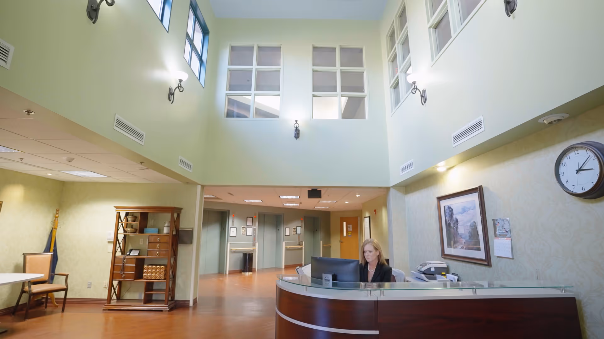 Reception area inside The Groves facility with a woman sitting behind a curved wooden desk with a computer. The space has high ceilings with windows near the top, wall-mounted lights, a clock, framed artwork, and a wooden shelving unit. There are doors and a hallway visible in the background.