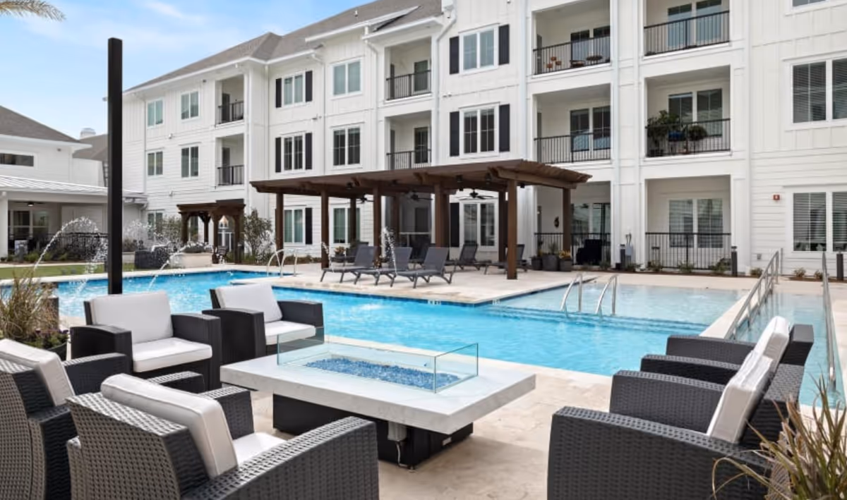 Outdoor swimming pool area at The Claiborne at Brickyard Crossing with lounge chairs, a fire pit surrounded by cushioned wicker chairs, and a wooden pergola. The background shows a three-story white building with balconies and large windows.