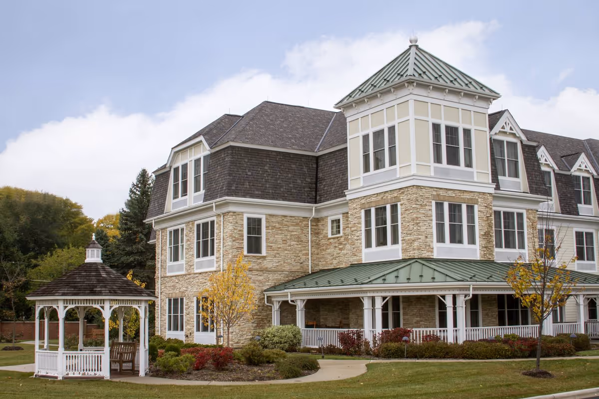 Exterior view of a multi-story senior living facility with stone and siding facade, multiple windows, and a green metal roof. In the foreground, there is a white gazebo with a shingled roof surrounded by landscaped bushes and trees with autumn foliage.