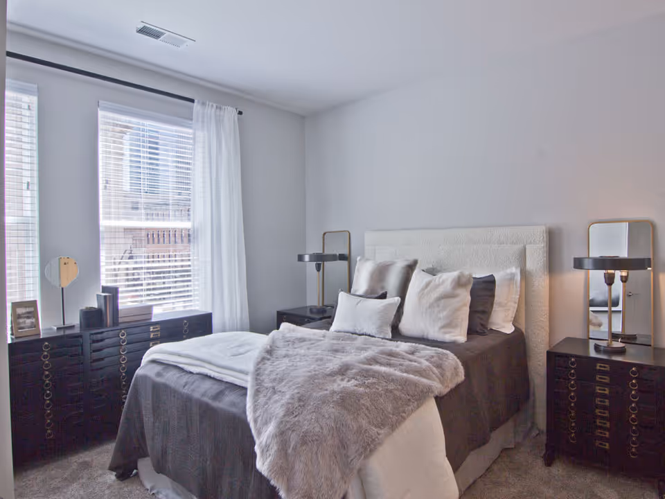 A neatly made bed with multiple pillows and a gray fur throw blanket in a bright bedroom. The room has two windows with white blinds and sheer curtains, two black nightstands with lamps and mirrors, and a black dresser with decorative items near the windows.