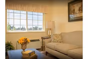 Cozy living room with a beige sofa, coffee table holding books and a vase of flowers, a side table with a lamp, and a large window.