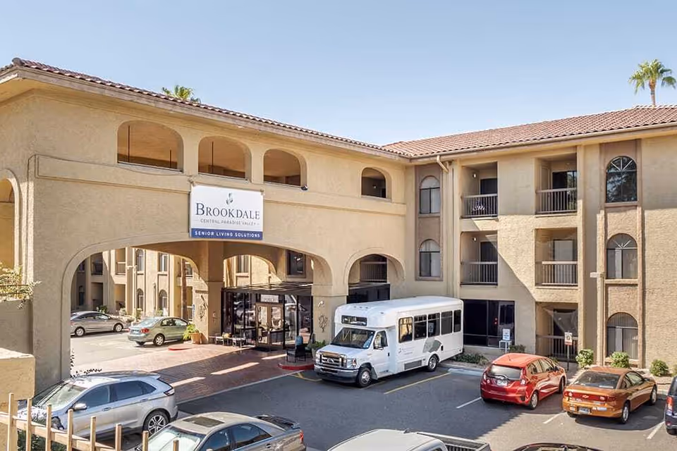 Exterior view of Brookdale Central Paradise Valley senior living facility showing a beige stucco building with arched openings, a covered entrance, several parked cars, and a white shuttle bus in the parking lot under a clear blue sky.