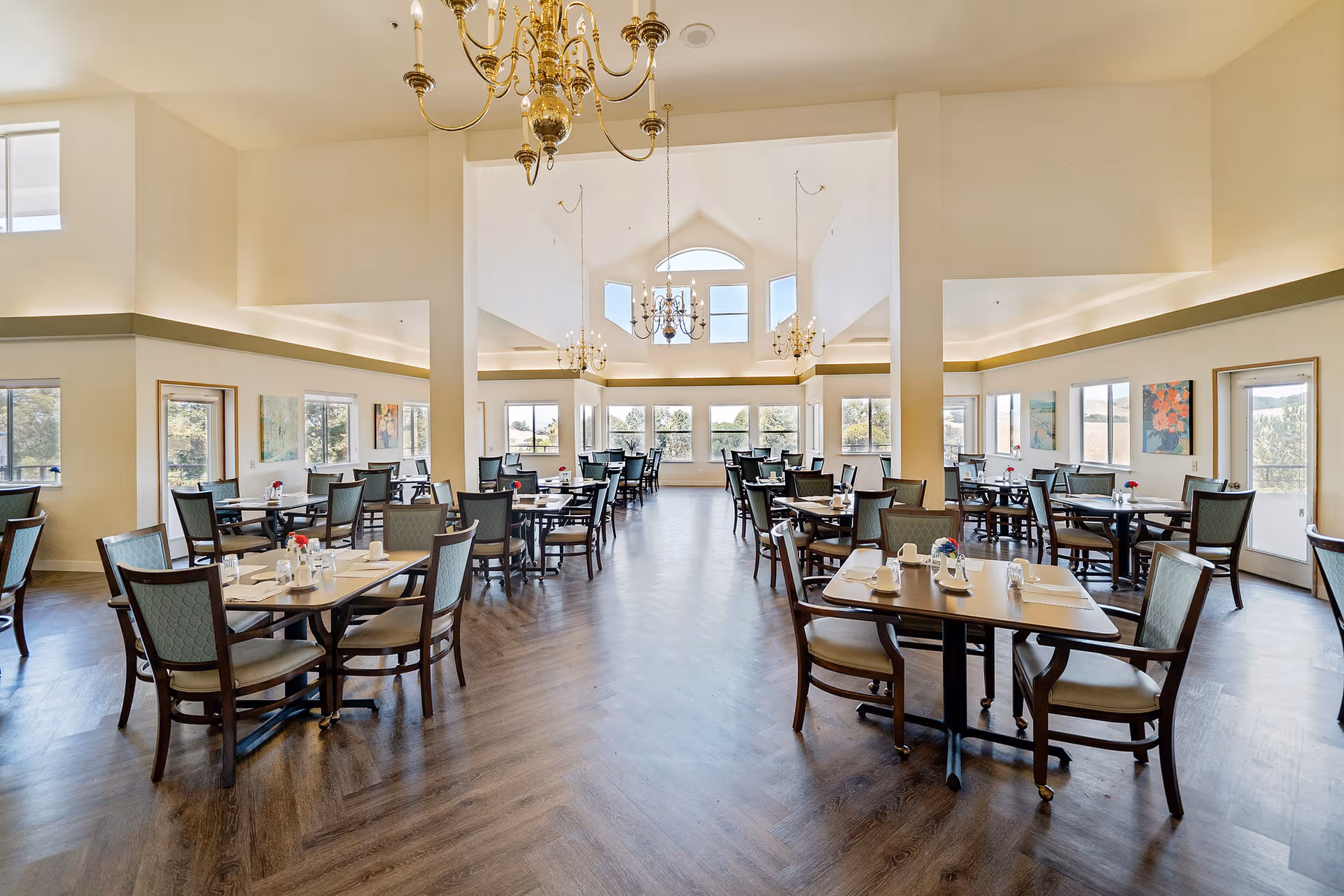 Spacious dining room with multiple tables and chairs arranged neatly. Each table is set with white cups, glasses, and small flower vases. The room features large windows allowing natural light to fill the space, high vaulted ceilings with elegant chandeliers, and light-colored walls adorned with colorful paintings.
