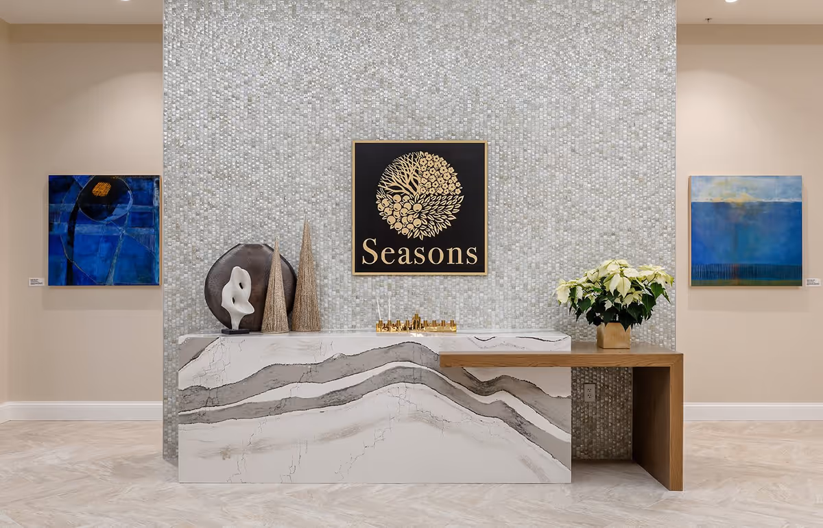 A modern reception desk with a marble-like front and a wooden side extension, decorated with abstract sculptures, two cone-shaped ornaments, and a potted white poinsettia plant. Behind the desk is a textured mosaic wall featuring a framed artwork with a gold floral design and the word 'Seasons'. On either side of the mosaic wall are two abstract paintings in shades of blue.
