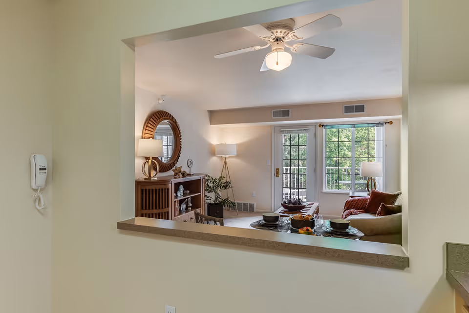 View through a kitchen pass-through into a furnished living room with a sofa, coffee table, lamps, ceiling fan, and glass door to a balcony.