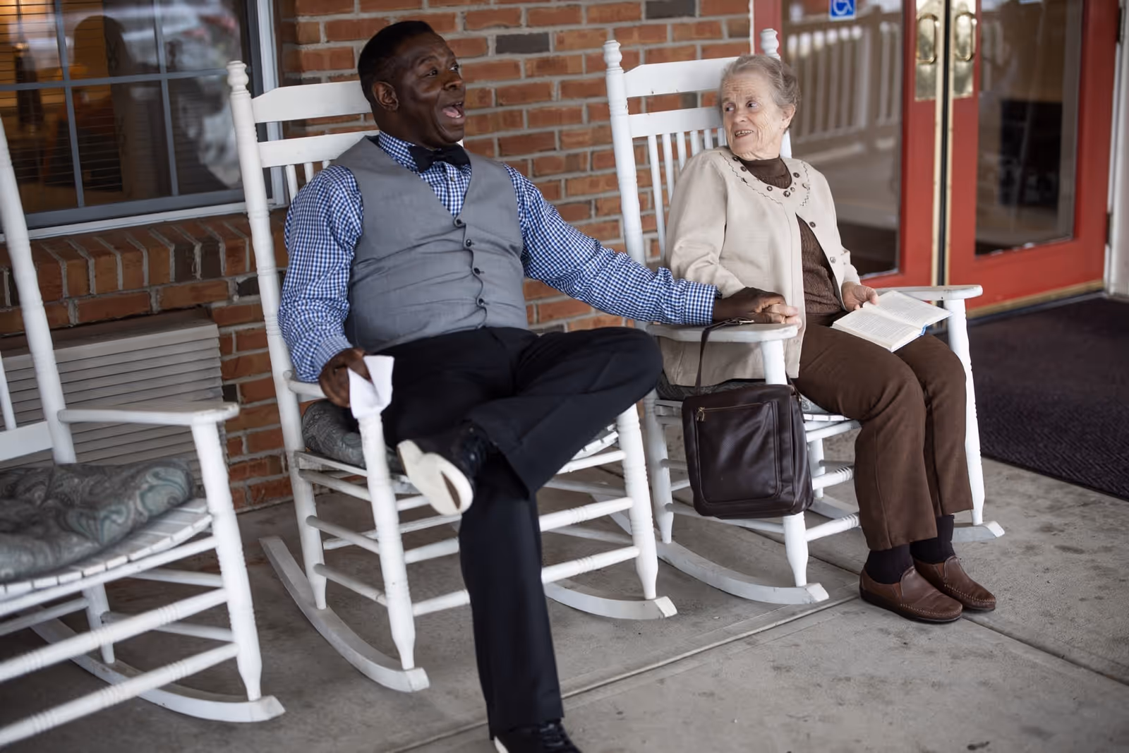 An elderly man and woman sitting on white rocking chairs on a porch outside a brick building. The man is wearing a gray vest, blue checkered shirt, and black pants, and is smiling while holding the woman's hand. The woman is wearing a beige cardigan, brown pants, and brown shoes, holding an open book on her lap. There is a brown bag hanging on the armrest of her chair and a red door in the background.