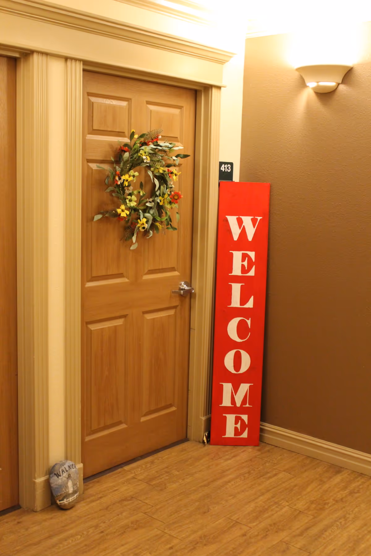 A hallway apartment door decorated with a floral wreath and a tall red vertical 'WELCOME' sign leaning beside it.