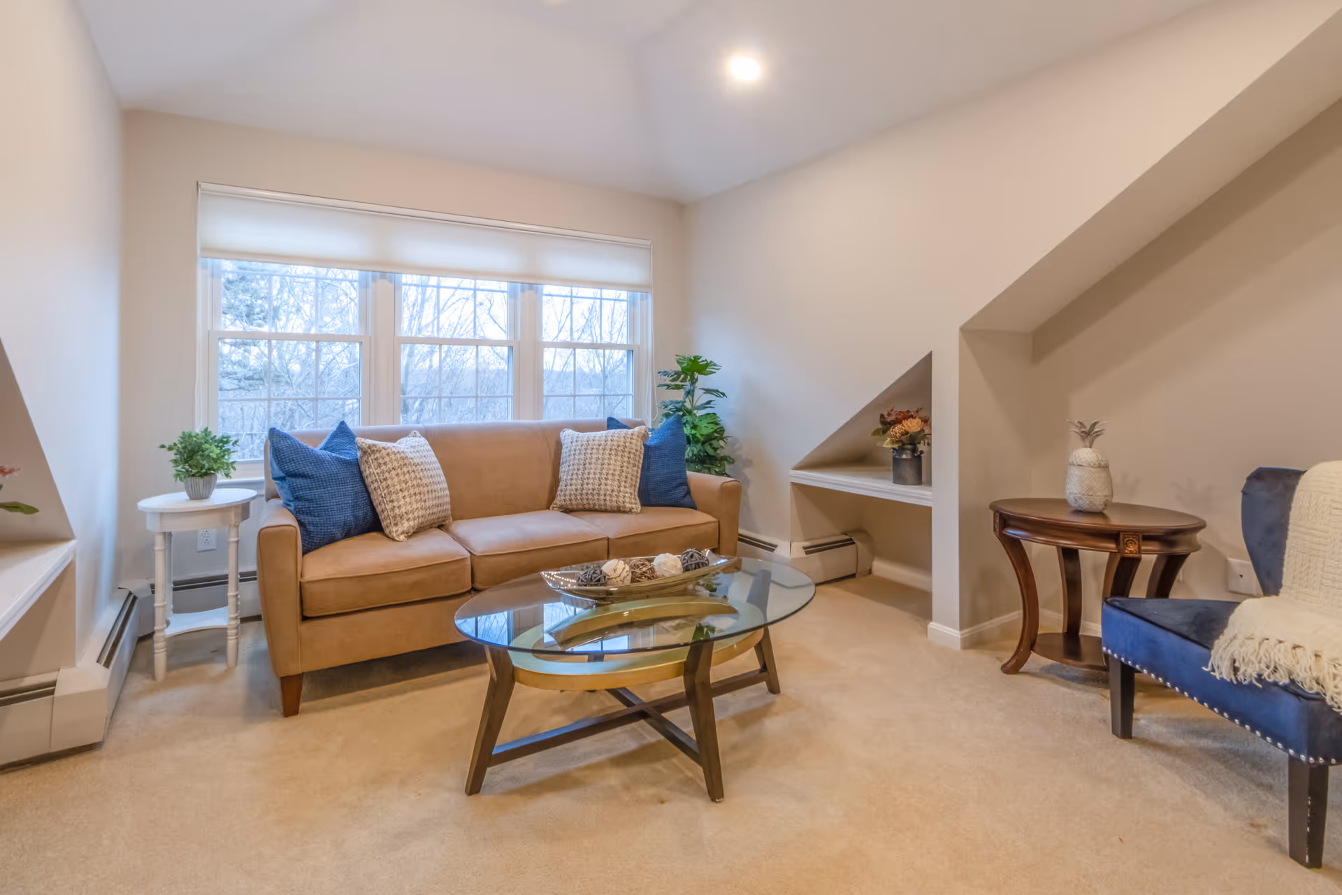 A cozy living room with a beige sofa adorned with blue and patterned throw pillows, a glass coffee table with decorative items, a blue upholstered chair with a white throw blanket, and two small side tables with plants and decorative objects. Large windows with white blinds allow natural light to fill the room.