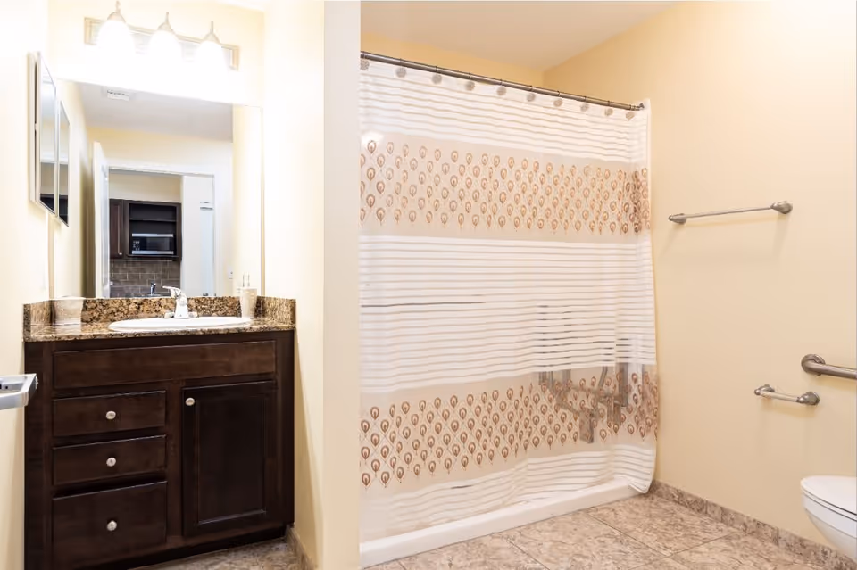 A bathroom with a dark wood vanity featuring a granite countertop and a sink. Above the vanity is a large mirror with three light fixtures. To the right, there is a bathtub with a shower curtain that has a pattern of brown and beige designs. The floor is tiled, and there are grab bars installed on the wall near the bathtub and toilet.