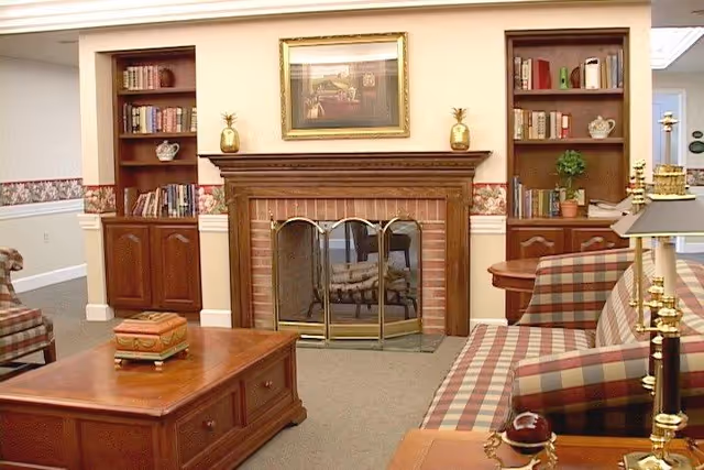 A cozy living room with a brick fireplace framed by wooden built-in bookshelves filled with books and decorative items. There is a wooden coffee table with a decorative box on it, a plaid upholstered sofa, and a brass lamp on a side table. The walls have a floral wallpaper border and a framed painting above the fireplace.